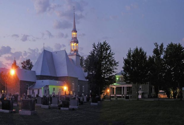 Photographie couleur prise de nuit, plan éloigné de l’arrière des bâtiments comprenant l’église, le presbytère et le cimetière, l’église est éclairée.