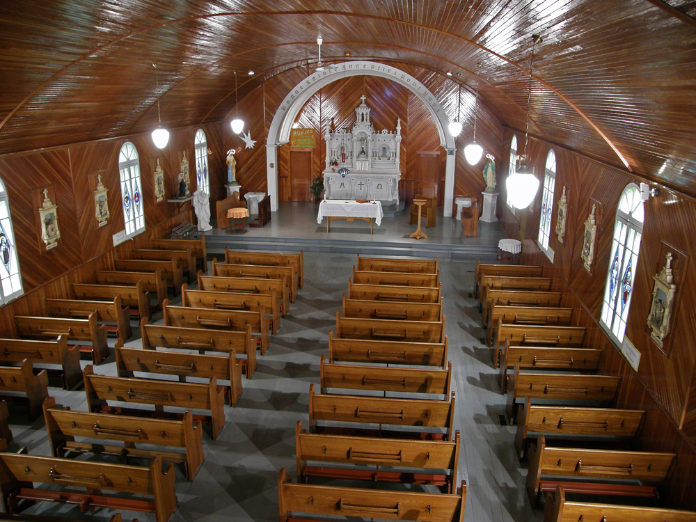 Photographie couleur, plan éloigné, intérieur d’une église dont le plafond et les murs sont en bois verni avec des fenêtres composées de vitraux colorés, la totalité du mobilier religieux est en place. 