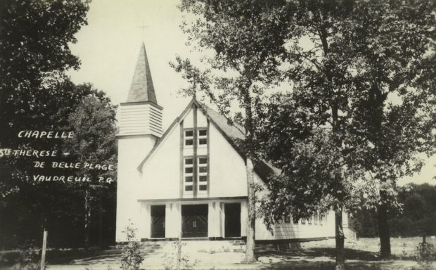 Photographie ancienne noir et blanc prise en été, plan rapproché, façade d’une église en bois peint en blanc, le clocher est au sommet d’une tour à la gauche de l’église.