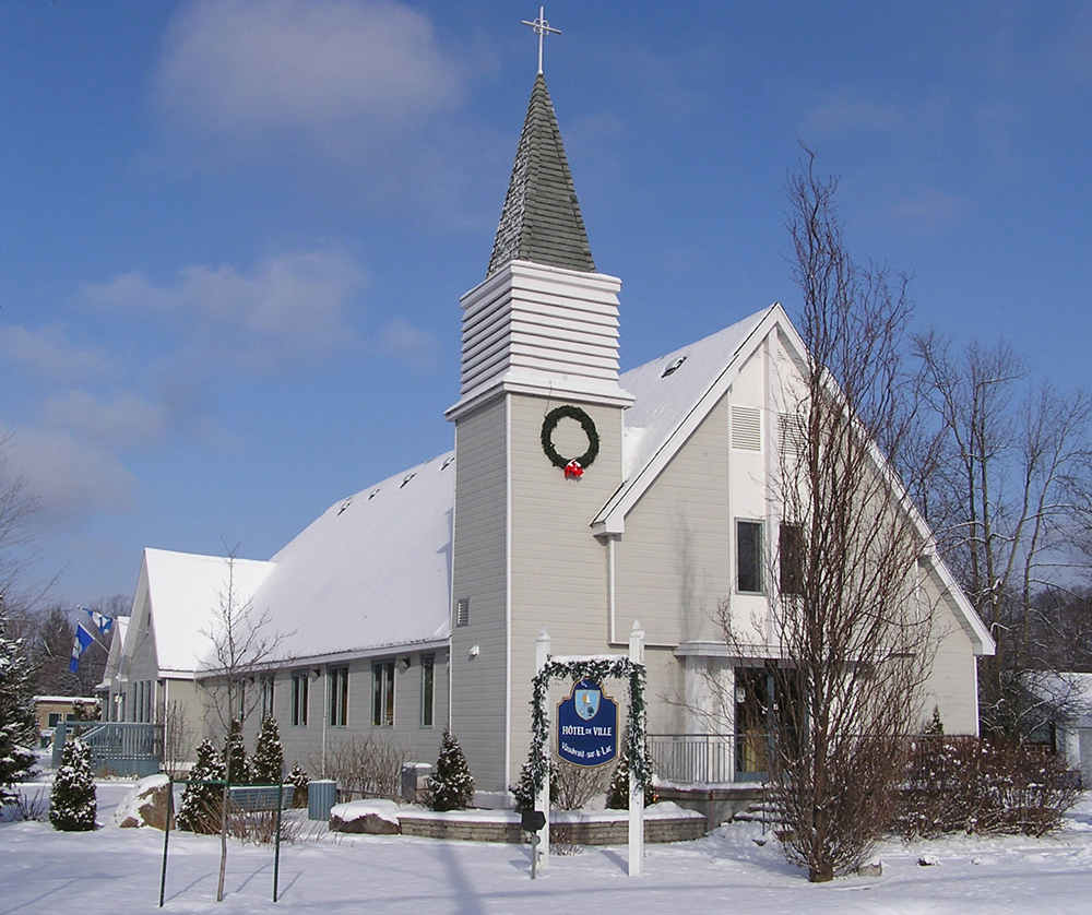 Photographie couleur prise en hiver, plan rapproché, église avec un clocher au sommet d’une tour à la gauche du bâtiment, devant l’église une affiche portant l’inscription Hôtel de ville .