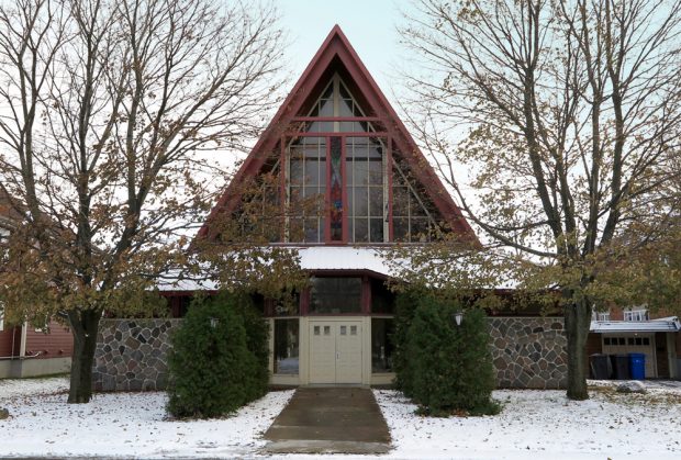 Photographie couleur prise en hiver, plan rapproché, façade d’une église dont le bas est en pierres et la toiture en pignon composée de vitraux colorés avec des arbres de chaque côté.  