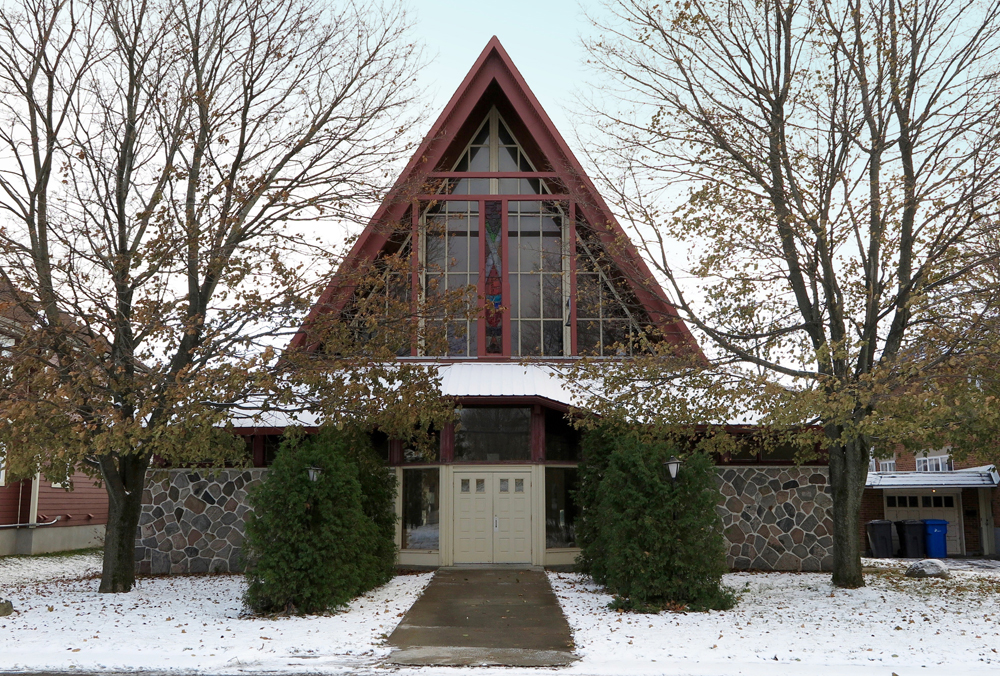 Photographie couleur prise en hiver, plan rapproché, façade d’une église dont le bas est en pierres et la toiture en pignon composée de vitraux colorés avec des arbres de chaque côté.  