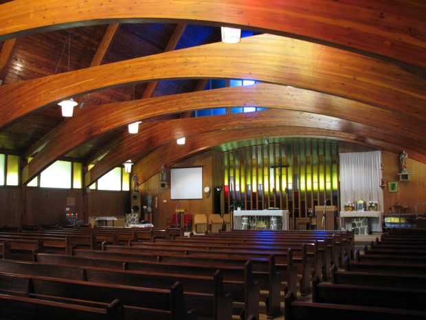 Photographie couleur, intérieur d’une église dont les poutres courbées et la charpente en bois verni sont apparentes.