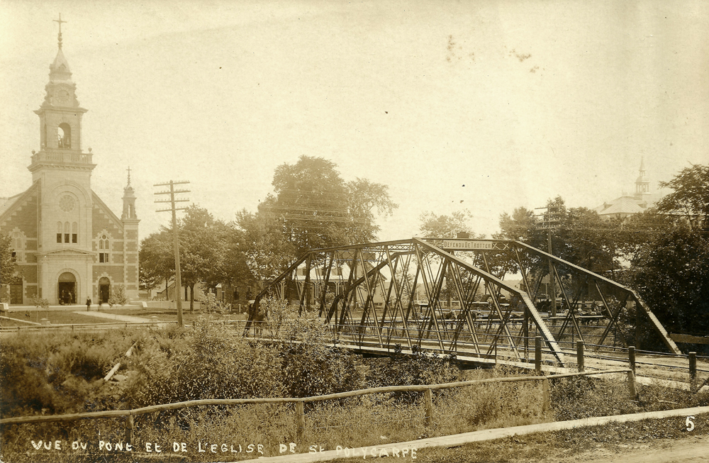 Photographie ancienne noir et blanc, plan éloigné, façade d’une église qui fait face à la route et à un pont de fer.