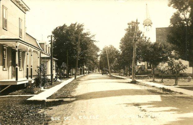 Photographie ancienne noir et blanc, plan éloigné, rue d’un village avec de chaque côté des trottoirs et des maisons, à l’arrière-plan, le clocher d’une église au-dessus des arbres.