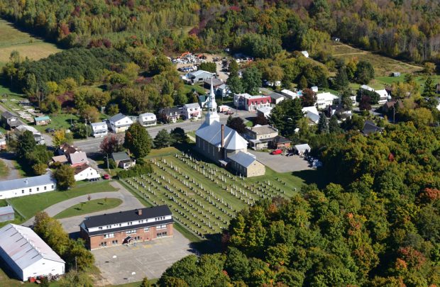 Photographie couleur, vue aérienne d’un village, au centre une église et un cimetière entourés de maisons et de bâtiments, autour la forêt.