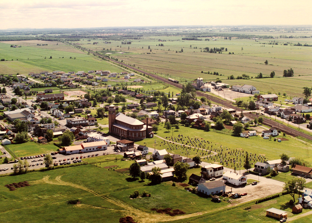 Photographie couleur, vue aérienne d’un village avec au centre une église, le village est entouré de terres agricoles.