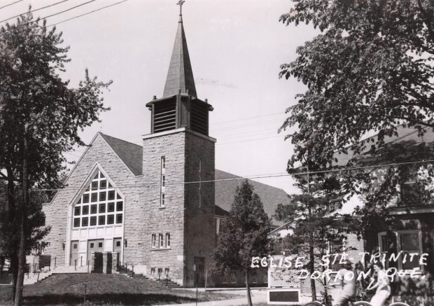 Photographie ancienne noir et blanc, façade d’un églises en pierres et d’un presbytère, en avant-plan à droite, deux jeunes filles en bicyclette.