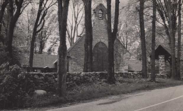 Photographie ancienne noir et blanc, façade d’une église en pierres entourée d’arbres, en avant-plan, un muret de pierres longe une route asphaltée.