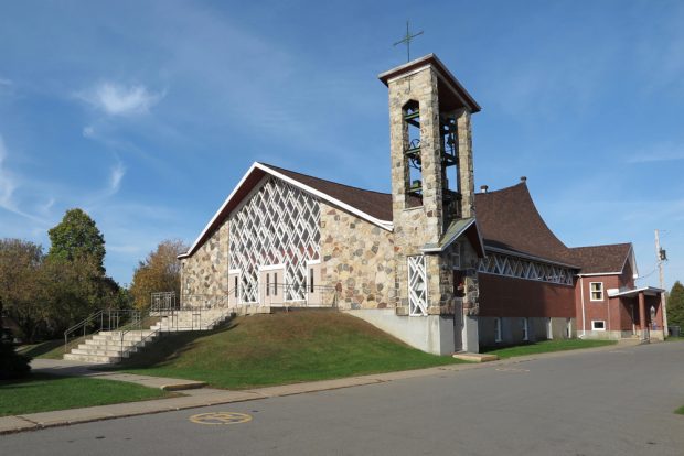 Photographie couleur, plan éloigné, église en pierres et en briques dont la façade comprend une verrière, à droite du bâtiment, un clocher carré en pierres dont les quatre côtés sont ouverts.