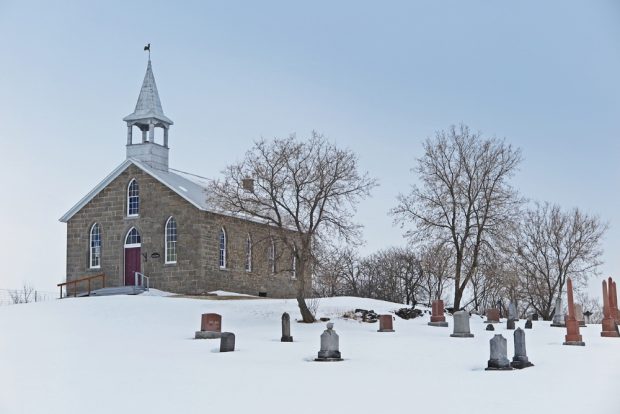Photographie couleur prise en hiver, plan éloigné, église de campagne en pierres vue de face, à droite un cimetière et des arbres.