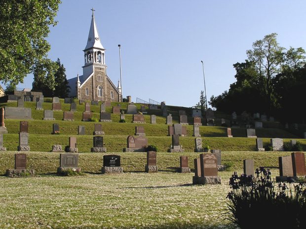 Photographie couleur, plan éloigné, à l’avant-plan, un cimetière construit en paliers sur une falaise, en arrière plan, la façade d’une église en pierres des champs et un clocher.