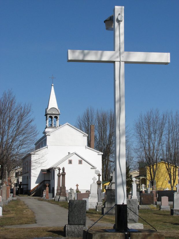 Photographie couleur, vue arrière d’une église, en avant-plan le cimetière et une grande croix blanche.
