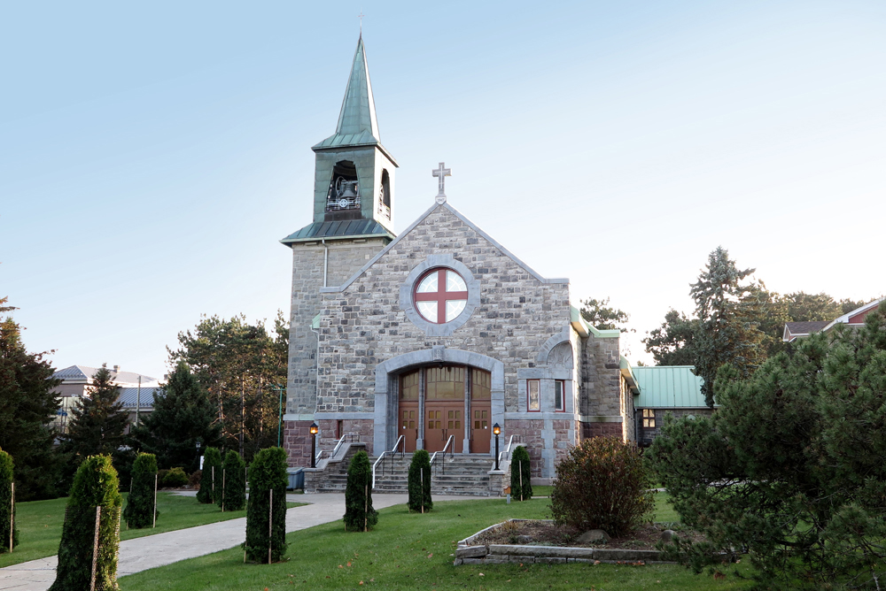 Photographie couleur, plan éloigné, façade d’une église en pierres surmontée d’une croix, à gauche, la tour d’un clocher en pierres taillées et sa toiture couleur cuivre oxydé, en avant-plan, un aménagement paysager.
