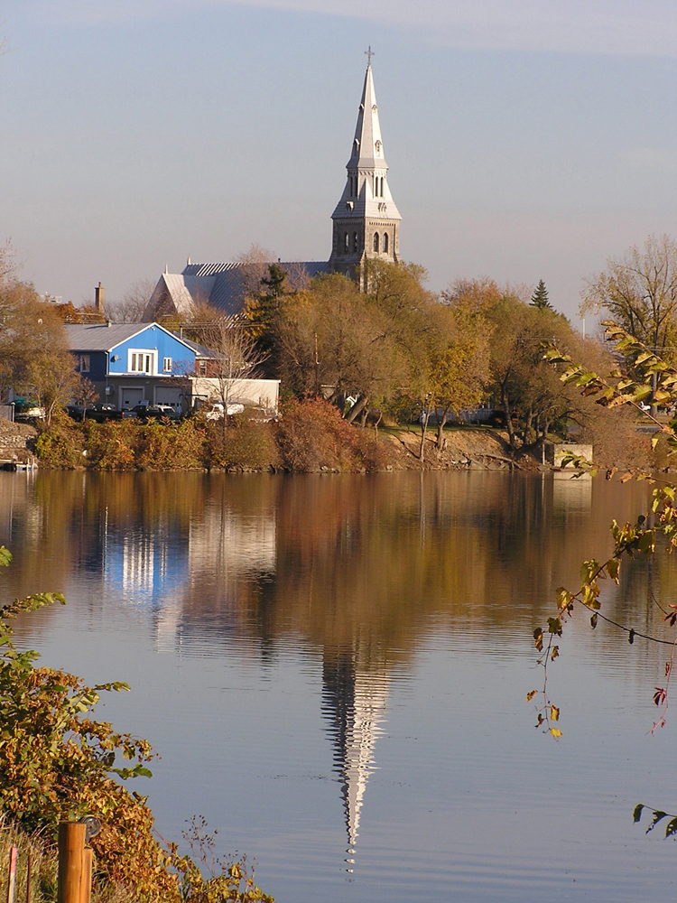 Photographie couleur, plan éloigné, clocher d’une église, d’une maison et d’arbres au bord de l’eau qui se reflètent sur l’eau.