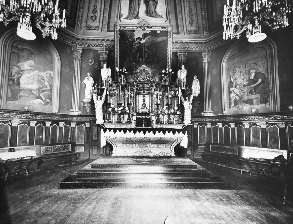 Photographie ancienne en noir et blanc, intérieur d’une église richement décorée comprenant du mobilier liturgique, des statues et des peintures religieuses.