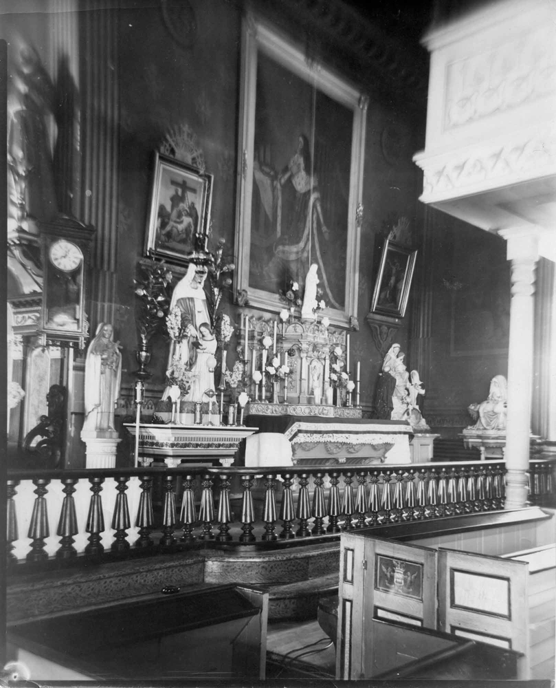 Photographie ancienne en noir et blanc,  intérieur d’une église, chapelle latérale richement décorée avec du mobilier liturgique, des statues et des peintures, en avant-blanc une balustrade et des bancs en bois vernis.