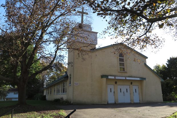 Photographie couleur, plan rapproché, façade d’une église en ciment couleur jaune, à gauche, un clocher carré surmonté d’une croix en bois blanc.