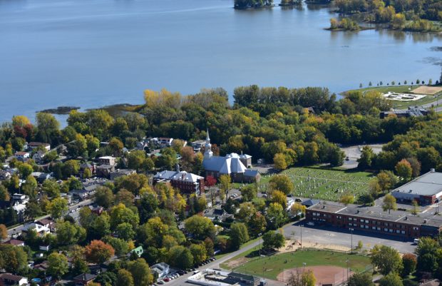 Photographie couleur, vue aérienne d’une ville au bord d’un grand lac, au centre, une église et un cimetière, à gauche des maisons et des arbres, à droite un parc et une école.