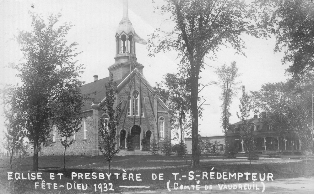 Photographie ancienne noir et blanc, façade d’une église et d’un presbytère avec des arbres, sur l’église des rubans descendent du clocher jusqu’au perron.