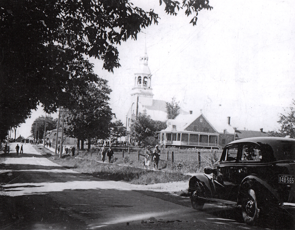 Photographie ancienne noir et blanc, en avant-plan, une route, une automobile et des personnes qui marchent sur un trottoir, en arrière-plan, un clocher d’église et un presbytère.