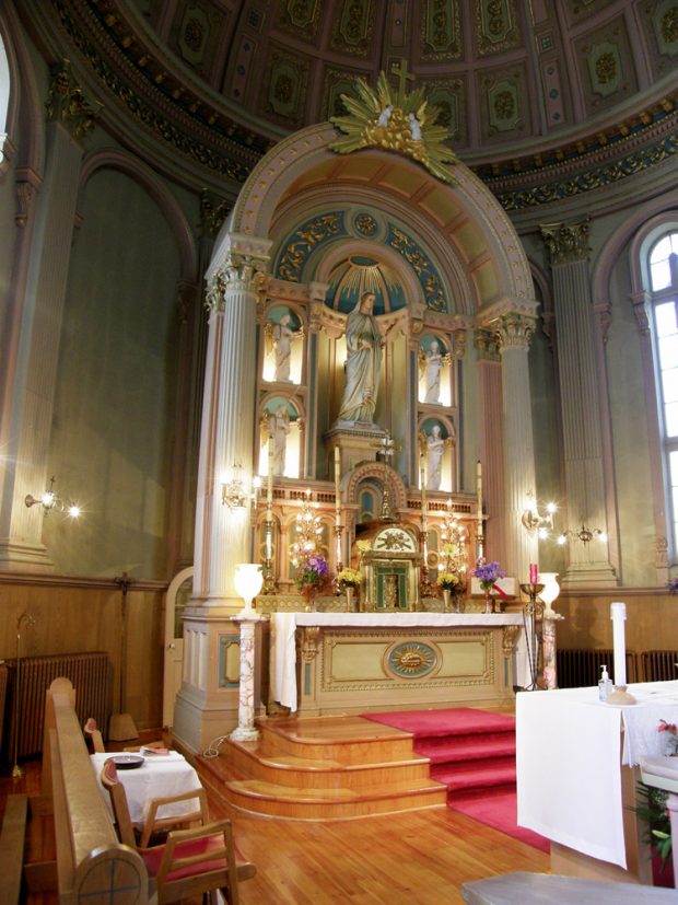 Photographie couleur, plan rapproché de l’intérieur d’une église avec du mobilier liturgique sur laquelle se trouve une grande statue entourée de quatre petites statuettes éclairées. 