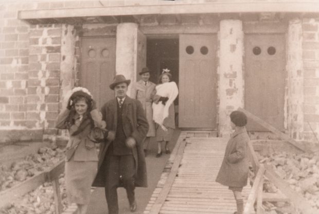 Photographie ancienne en noir et blanc, un enfant et deux couples, dont une femme qui tient un bébé dans ses bras, sortent par les portes avant d’une église en construction.  