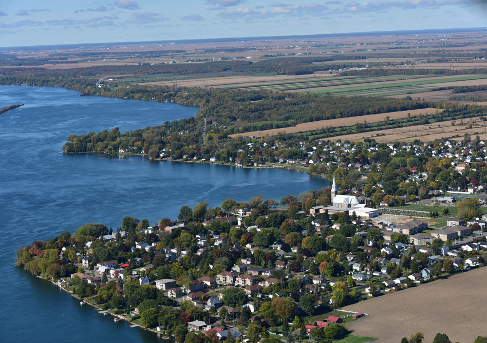Photographie couleur, vue aérienne d’une agglomération urbaine sur les rives d’un grand cours d’eau, au centre, l’église et son clocher, en arrière-plan, de grandes terres agricoles.