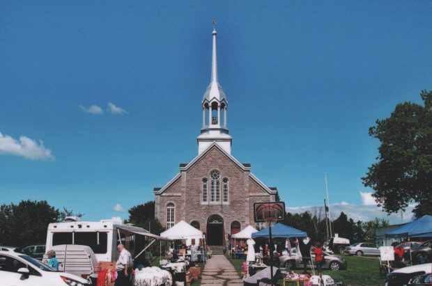 Photographie couleur, façade d’une église devant laquelle se trouvent des personnes, des automobiles, des tables et des chapiteaux.