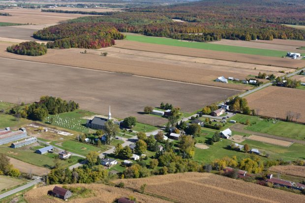 Photographie couleur, vue aérienne du rang principal d’un village, au centre l’église, le  cimetière et des maisons entourés de grandes terres agricoles. 