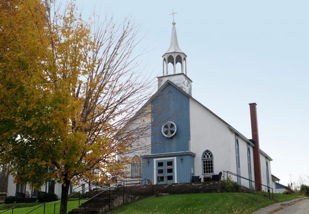 Photographie couleur, façade d’une église en crépi et en bois peinte en blanc et bleu, le clocher est sans cloche, sur le perron un mobilier de jardin et un chien.