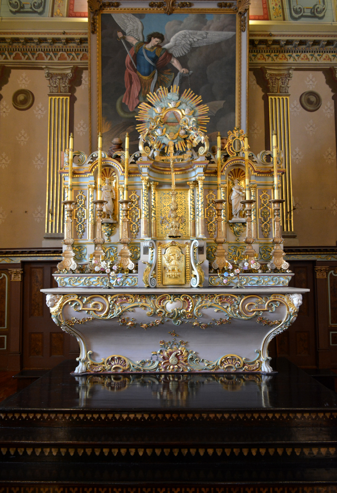 Photographie couleur, plan rapproché, mobilier liturgique richement sculpté et décoré à la feuille d’or, composé d’un tombeau et d’une table surmontée d’une partie supérieure sur laquelle est déposée deux statues et des chandeliers en bois et feuilles d’or. 