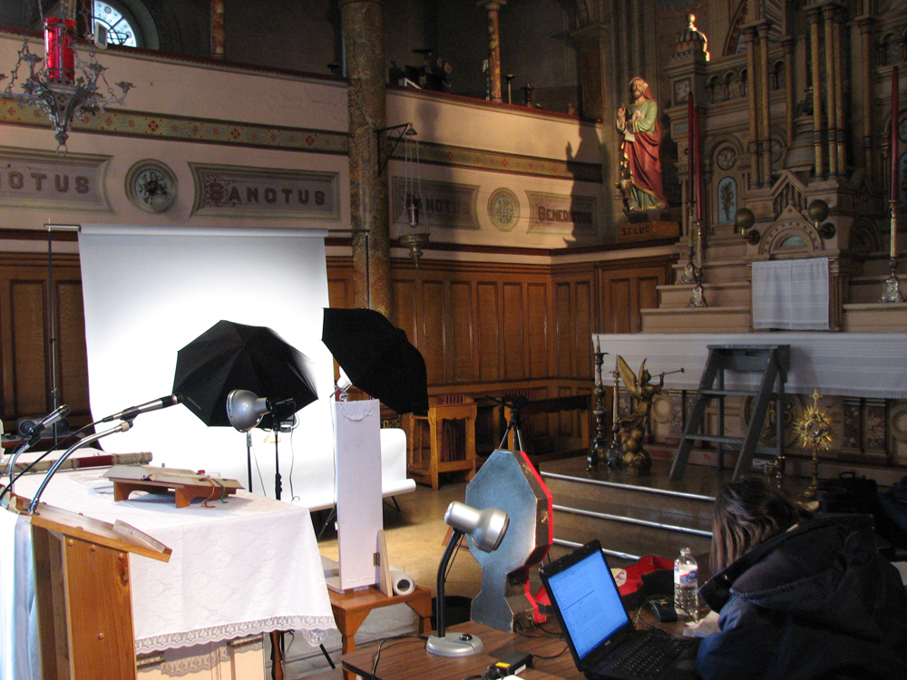 Photographie couleur, plan rapproché, intérieur d’une église, du matériel photographique et différents objets religieux sont dispersés sur des tables, une femme travaille devant un ordinateur.  