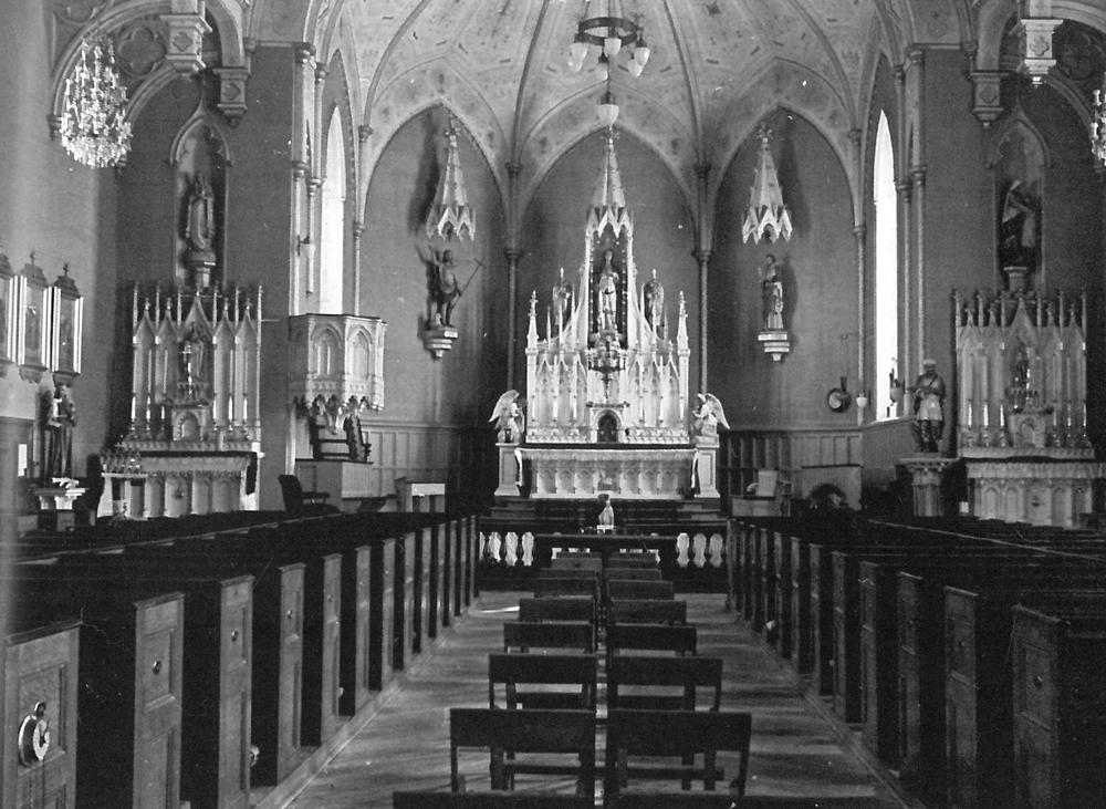 Photographie ancienne en noir et blanc, plan éloigné, intérieur d’une église, en arrière-plan, du mobilier liturgique et plusieurs statues, en avant-plan, plusieurs rangées de bancs en bois.