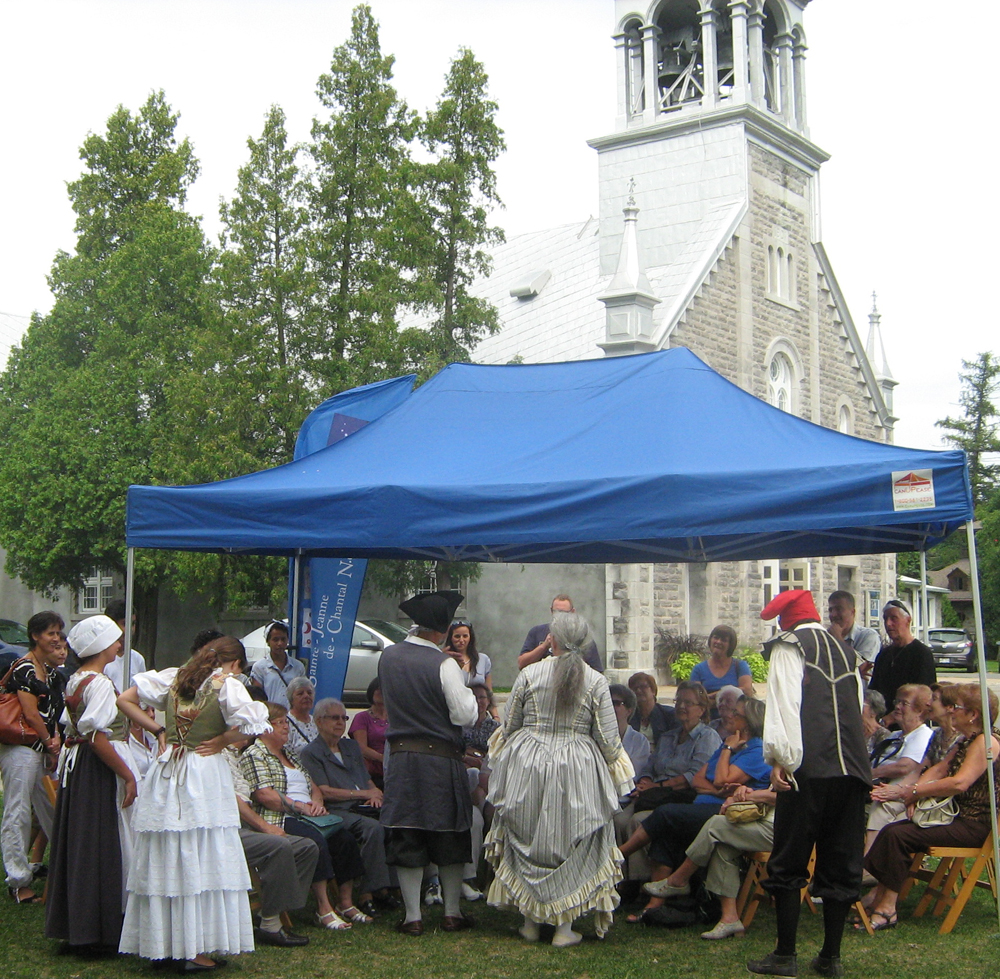 Photographie couleur, plan rapproché, sous un chapiteau situé entre une chapelle et une église en pierres, des comédiens costumés et des adultes participent à une activité culturelle.