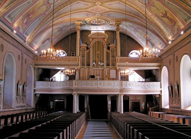 Photographie couleur, plan éloigné, intérieur d’une église dont la voûte est décorée, à l’arrière-plan, une tribune et un grand orgue à tuyaux en cuivre, en avant-plan, des rangées de bancs en bois. 