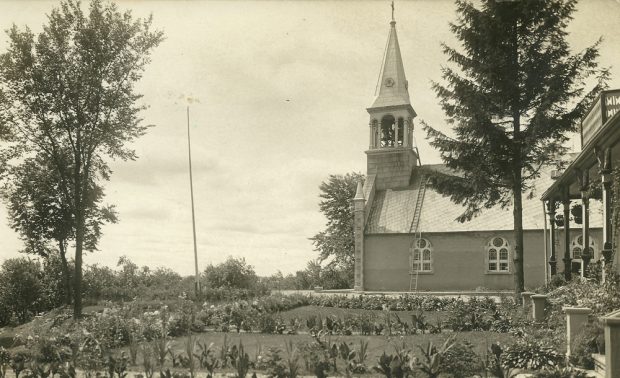 Photographie ancienne en noir et blanc, vue sur un grand jardin fleuri à l'avant d'un bâtiment, en arrière-plan, vue latérale d'une église et du clocher.