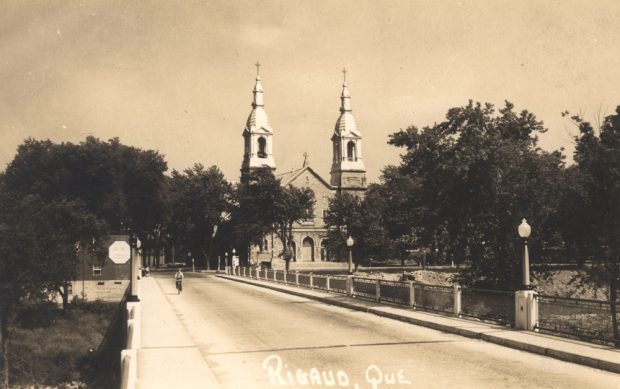 Photographie ancienne noir et blanc, un cycliste sur un large pont pavé qui même à une grande église surmontée de deux clochers et entourée d’arbres.