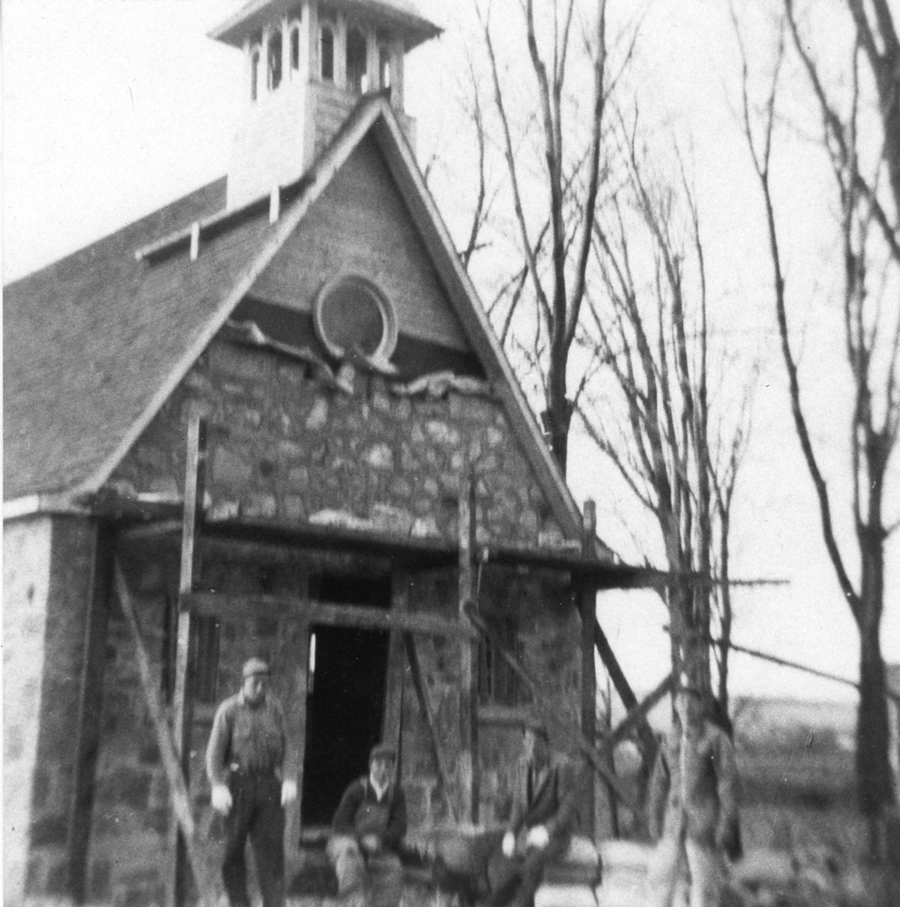 Photographie ancienne en noir et blanc, quatre travailleurs devant la façade d’une petite chapelle en pierres des champs en construction avec des échafaudages. 