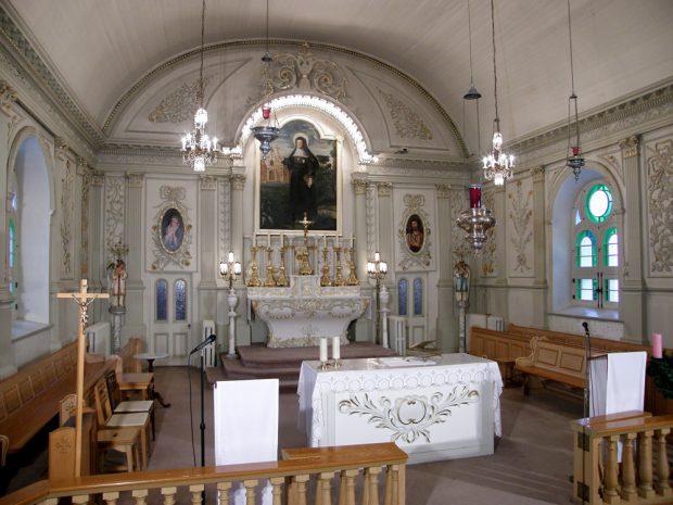 Photographie couleur, plan éloigné, intérieur d’une église dont la voûte et les murs sont entièrement en lattes de bois peint en blanc, à l’arrière plan, du mobilier liturgique et des œuvres d’art religieux, à l’avant-plan du mobilier religieux et une balustrade en bois verni.