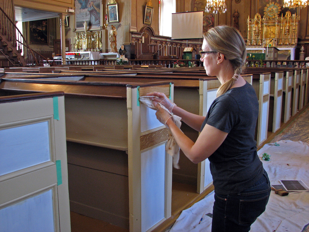 Photographie couleur, plan rapproché, une femme à genoux, munie de gants, travaille à l’aide d’un morceau de tissu à la restauration d’un banc en bois à l’intérieur d’une église richement décorée.