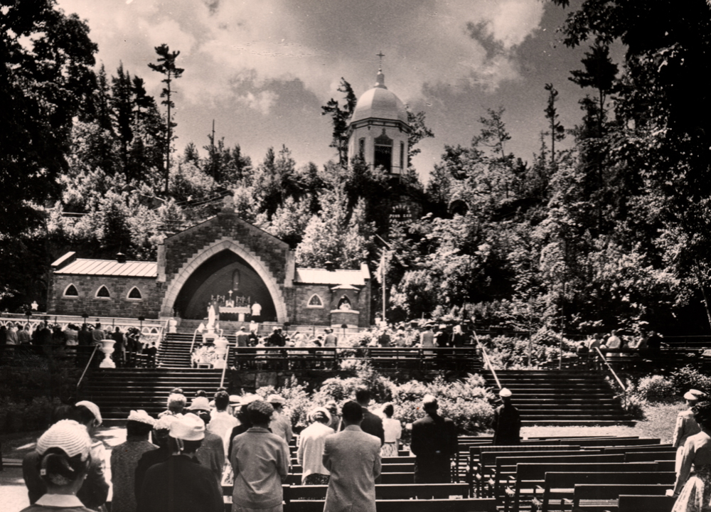 Photographie ancienne en noir et blanc, plan éloigné, en avant-plan, des pèlerins se tiennent debout devant leurs bancs pour une cérémonie religieuse célébrée en plein air, en arrière plan, une chapelle à aire ouverte au milieu d’une colline surmontée d’une rotonde et entourée d’arbres.