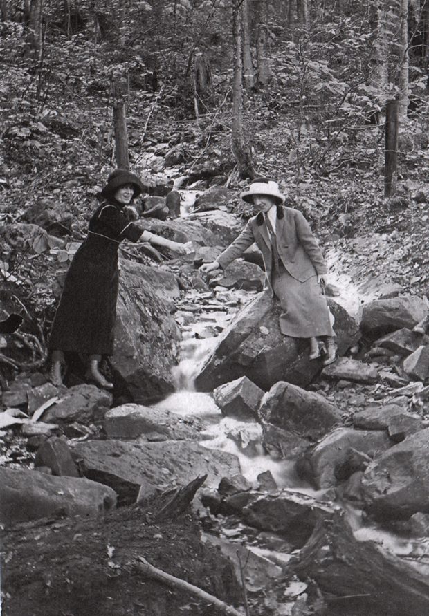 Photographie ancienne en noir et blanc, plan rapproché, deux jeunes femmes avec des tasses dans les mains puisent l’eau d’un ruisseau au milieu de rochers. 