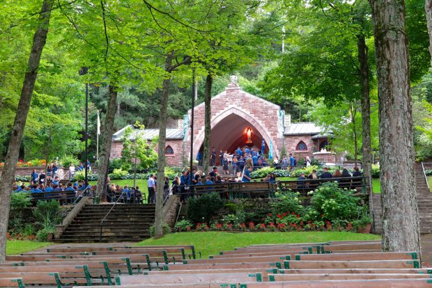 Photographie couleur, plan éloigné, une foule est rassemblée à l’extérieur près d’une chapelle à aire ouverte entourée d’arbres et de jardins. 	