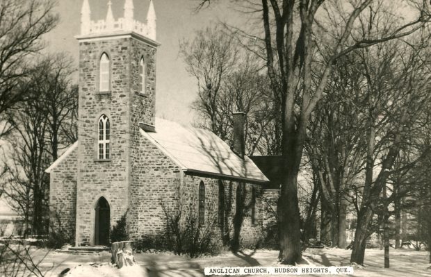 Photographie ancienne en noir et blanc, prise en hiver, plan éloigné, façade et vue latérale d’une église en pierres des champs, avec son clocher carré surmonté de pinacles, entourée d’arbres.