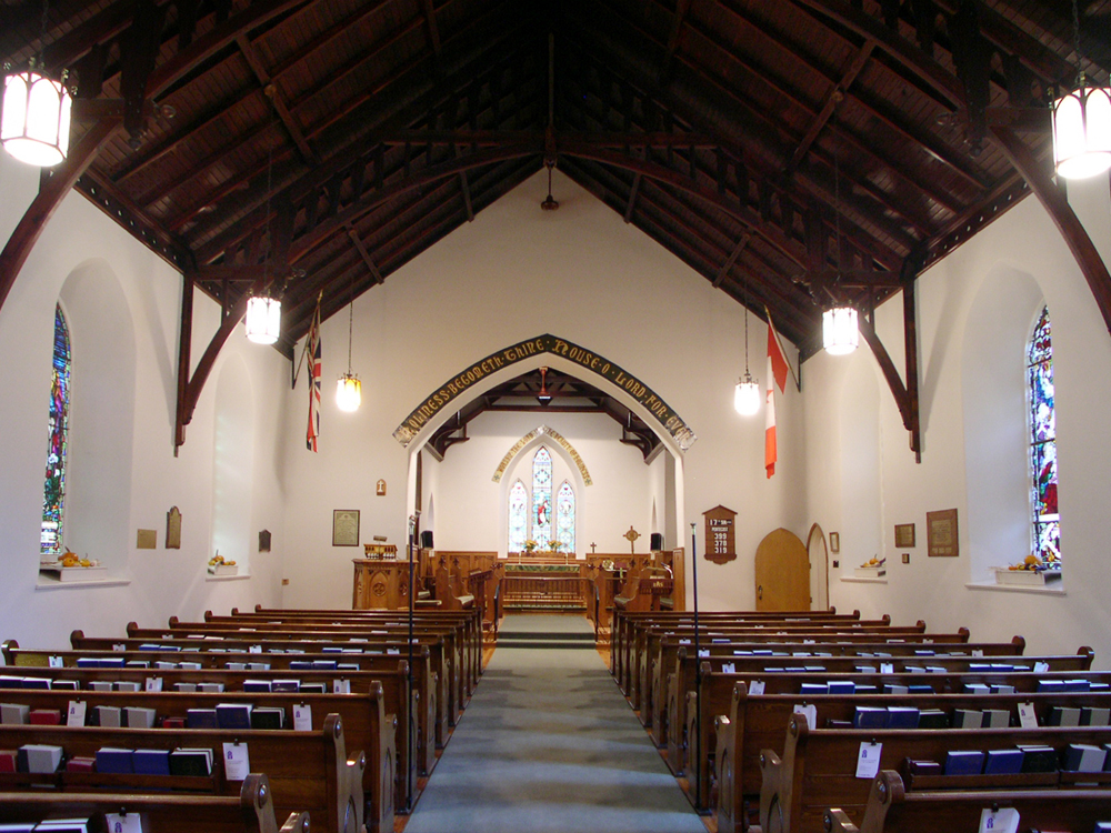 Photographie couleur, plan éloigné, intérieur d’une église dont la voûte est entièrement en bois sculpté, en avant-plan, des bancs en bois de chaque côté d’une allée, sur les côtés des vitraux colorés, en arrière-plan, une verrière de vitraux colorés. 