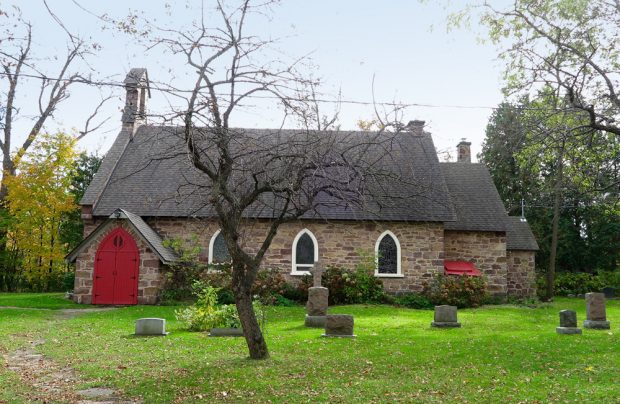 Photographie couleur, plan éloigné, vue latérale d’une église en pierres des champs entourée d’arbres, en avant-plan, des pierres tombales.