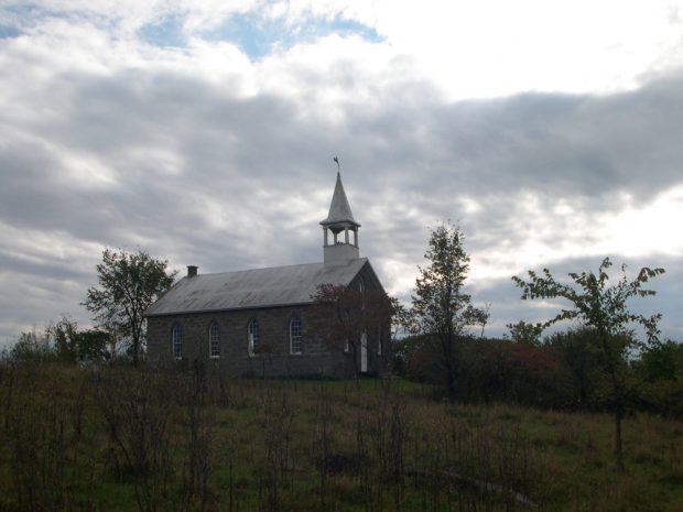 Photographie couleur, plan éloigné, vue latérale d’une petite église de campagne en pierres des champs entourée d’arbres sous un ciel nuageux.