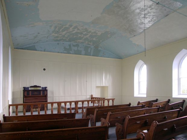 Photographie couleur, intérieur d’une église dont les murs sont blancs et sans décoration avec deux fenêtres, en avant-plan, des bancs en bois, en arrière-plan un petit harmonium entouré d’une balustrade en bois.  