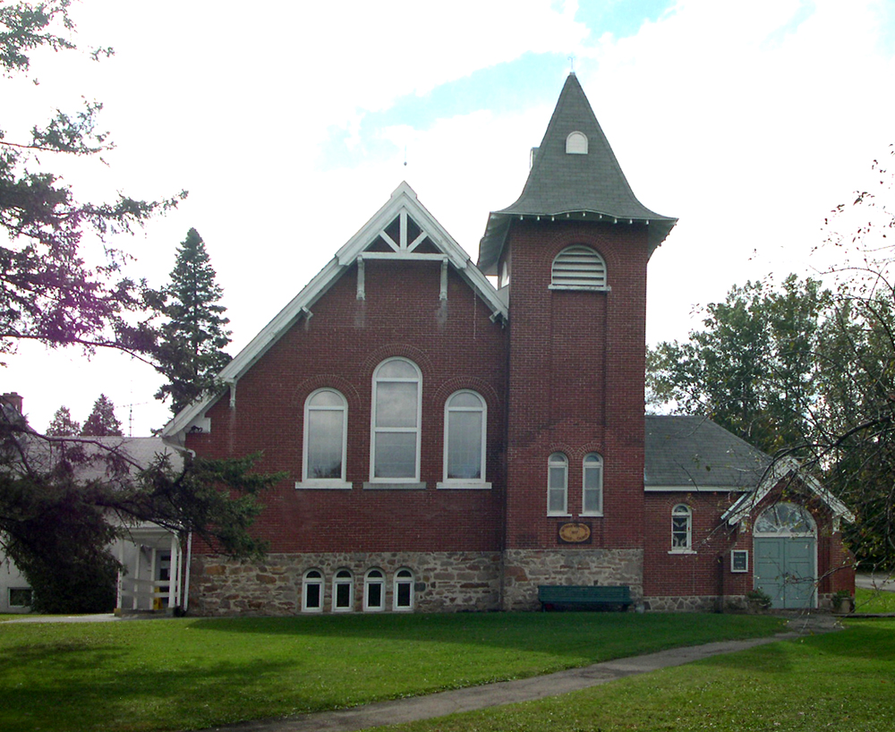 Photographie couleur, plan éloigné, façade d’une église en briques rouges avec un toit en pente prononcé et un clocher carré, à l’avant, un trottoir mène à une porte d’entrée en bois à droite du bâtiment.